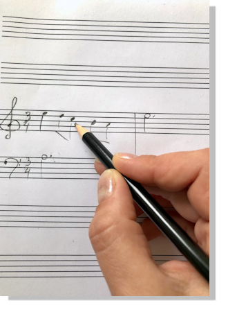 Eine Hand hält einen Bleistift und schreibt einen Violin- und einen Basschlüssel sowie einzelne Noten auf Notenpapier. 